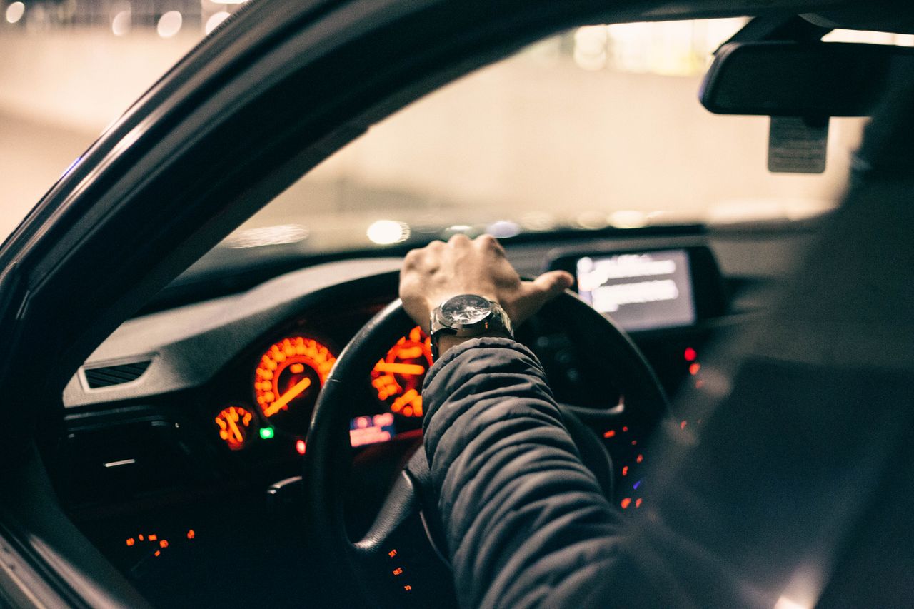 A puffer-jacketed driver grips the steering wheel, dashboard glowing with illuminated gauges.