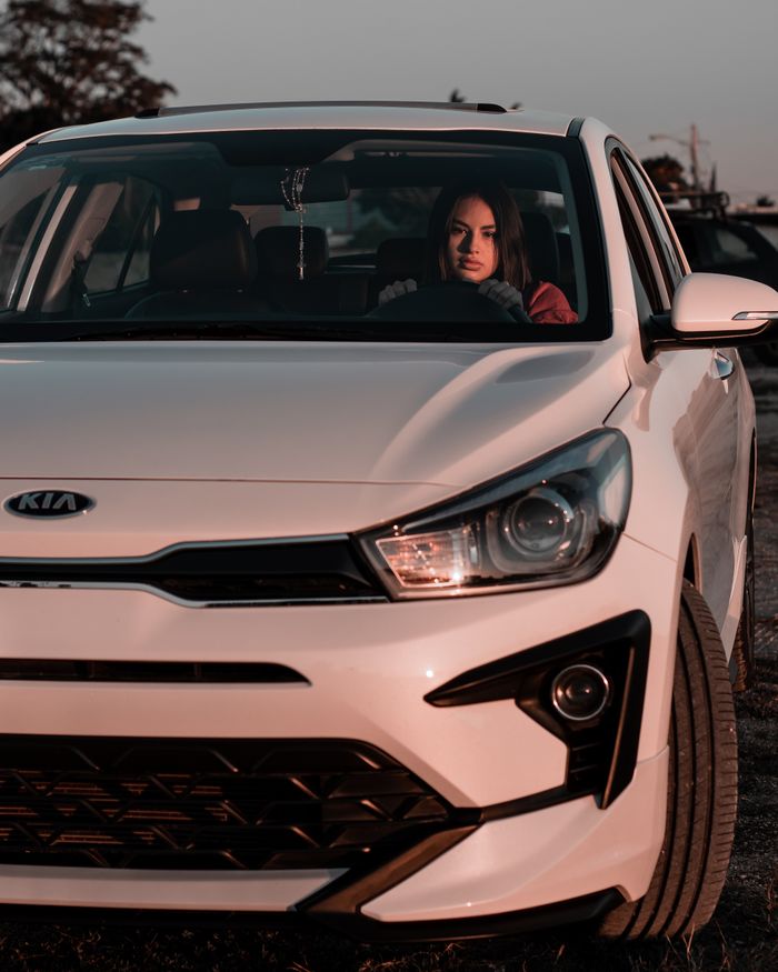 A woman sitting in a white Kia car, captured in warm evening light outdoors.
