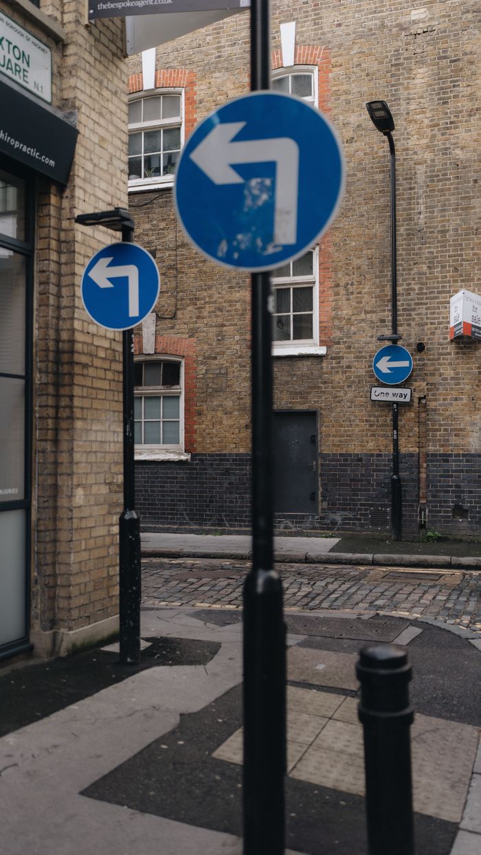A city alley features multiple blue directional road signs pointing left, surrounded by brick buildings.