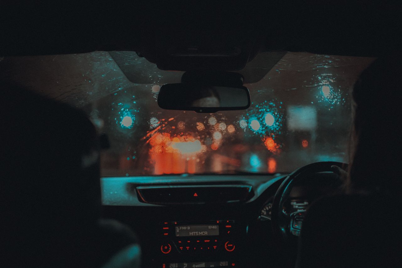 A cinematic view from inside a car driving through rainy Manchester at night with neon lights.